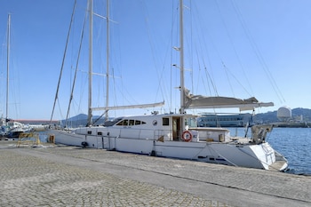 Vista lateral de un gran velero blanco de dos mástiles amarrado en un muelle adoquinado. El cielo es azul claro, el agua tranquila.