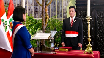 Hombre en traje negro con corbata roja y banda bicolor, de pie frente a mujer de espaldas en vestido azul con banda similar. Incluye banderas peruanas, crucifijo, atril