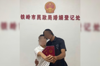 Una pareja sostiene dos certificados de matrimonio rojos frente a una pared blanca con caracteres chinos y un escudo oficial dorado