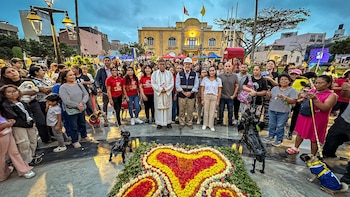 Multitud de personas y perros reunidos en una plaza frente a un arreglo floral en forma de corazón y un edificio público
