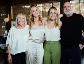 Cuatro personas sonrientes, tres mujeres y un hombre con barba, posan juntas en un interior con persianas de madera al fondo