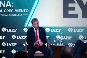 Luis Caputo viste traje oscuro y corbata roja, sentado en un sillón de madera en un escenario con el logotipo del IAEF al fondo