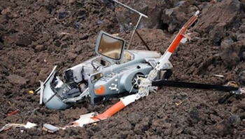 Helicóptero gris destrozado yace sobre un terreno rocoso y oscuro. La puerta de la cabina está abierta, las palas del rotor principal rotas y el fuselaje dañado