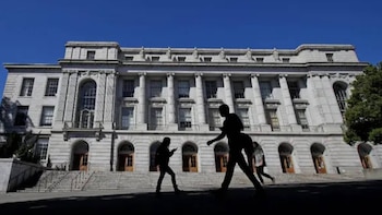 Siluetas de tres personas caminan frente a un gran edificio de arquitectura clásica de piedra clara, con columnas, ventanas altas, puertas arqueadas y amplias escalinatas bajo un cielo azul