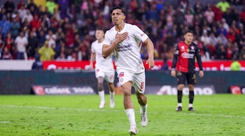 Un futbolista con uniforme blanco corre sobre el césped con la mano en el pecho ante la tribuna y otros jugadores.