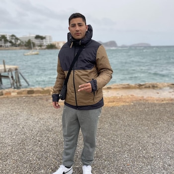 Un hombre joven con una chaqueta marrón y negra, pantalón gris y zapatillas blancas posa de pie frente al mar