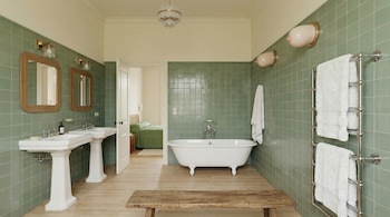 Baño con paredes verdes, dos lavabos de pedestal, bañera blanca exenta, suelo de madera clara y toallero cromado con toallas.