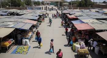 Vista aérea de un mercado bullicioso con puestos cubiertos por toldos, personas caminando y comprando, y un arco con la palabra 'ECUADOR' al fondo.