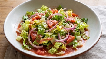 Ensaladera blanca con lechuga troceada, trozos de tomate, aros de cebolla morada y un líquido rojizo sobre una mesa de madera.