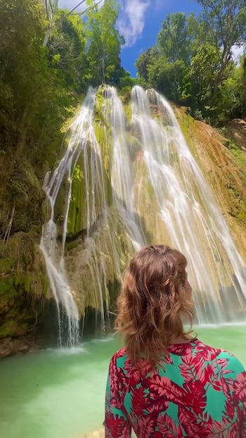 Una mujer de espaldas con ropa de estampado tropical observa una cascada alta que cae sobre un estanque de agua verde rodeado de árboles