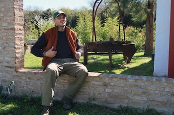 Un hombre con gorra y chaleco sentado sobre un muro de ladrillos en un jardín rodeado de árboles y vegetación