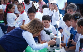 Una mujer maniobra un microscopio en una mesa frente a varios niños con uniformes escolares y letreros de Clubes de Ciencias