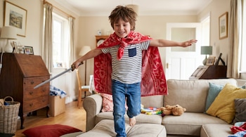 Niño sonriente con camiseta a rayas y jeans, usando una toalla roja como capa y sosteniendo una espada de juguete, salta en un puf de una sala de estar.