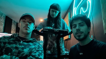 Tres hombres en un estudio de música con luces de neón azul. Uno lleva gorra y chaqueta de camuflaje, otro tiene pelo largo y sostiene un teclado, y el tercero una camiseta negra.