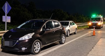 Dos automóviles estacionados en la banquina de una carretera nocturna junto a un cono de tránsito y una ambulancia con luces encendidas