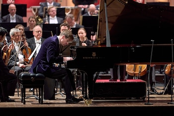 Un hombre de traje oscuro toca un piano de cola negro ante músicos con instrumentos de cuerda en un escenario iluminado