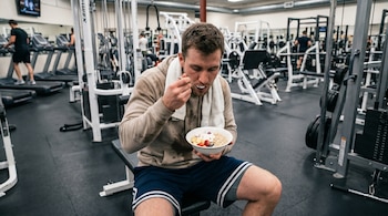 Un hombre en un gimnasio come yogur, avena y frutas de un bol blanco mientras está sentado en un banco. Viste sudadera beige y shorts. Máquinas de ejercicio al fondo.