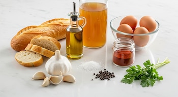 Ingredientes de cocina sobre encimera blanca: pan, ajos, aceite de oliva, caldo, huevos, pimentón, sal, pimienta y perejil fresco. Sin etiquetas ni logotipos.