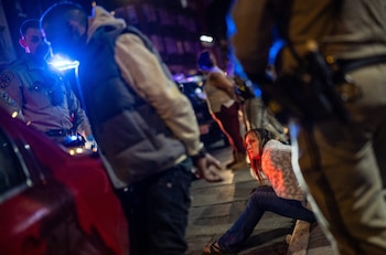 Vista nocturna de agentes de policía, una persona esposada de espaldas, y una mujer sentada en el suelo, con luces de emergencia azules y rojas