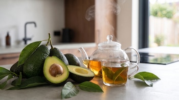 Aguacates enteros y partidos, con hojas verdes, junto a una tetera de cristal y una taza con té de hojas humeante en una encimera de cocina.