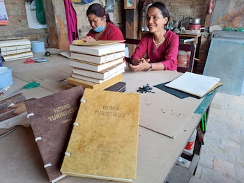 Dos mujeres en un taller, una sonríe y la otra trabaja en libros apilados. En primer plano, dos libros con cubiertas marrones y texto 'Homo Turisticus'