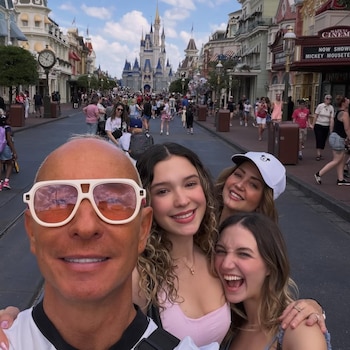 Erik Rubín en primer plano, sonriendo con gafas de sol, junto a Andrea Legarreta y sus dos hijas, en un selfie con el Castillo de Cenicienta al fondo