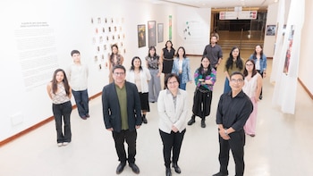 Un grupo de catorce personas posa de pie en el pasillo de una galería de arte con obras y textos explicativos en las paredes