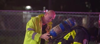 Un hombre con chaqueta amarilla reflectante sostiene el cilindro de aire en la espalda de un bombero inclinado por la noche