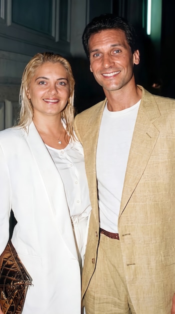 Un hombre con traje beige y camiseta blanca posa sonriendo al lado de una mujer rubia vestida con chaqueta y blusa blancas.