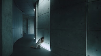 Una persona vestida con ropa clara permanece sentada en el suelo de un pasillo de hormigón junto a una franja de luz en la base de la pared.