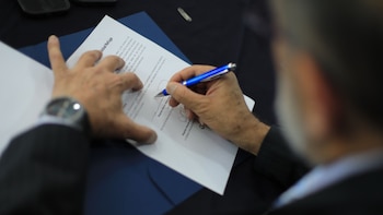 Primer plano de una mano escribiendo con un bolígrafo azul sobre un documento blanco. La mano lleva un reloj y el papel descansa sobre una superficie oscura