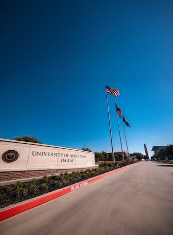 Letrero de la Universidad de North Texas en Dallas con banderas (EE.UU., Texas, UNT Dallas) y una carretera de acceso con acera roja bajo un cielo azul despejado