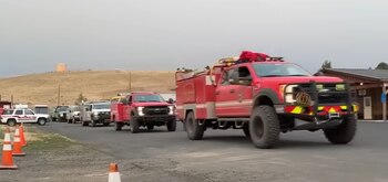 Un convoy de camiones de bomberos y vehículos de emergencia avanza por una carretera de tierra. Hay conos de tráfico, un edificio y un depósito de agua al fondo