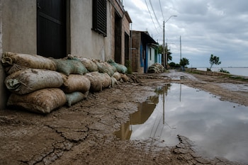 Sacos de arena apilados junto a la pared de una casa en un camino de barro con un gran charco y un cuerpo de agua al fondo.