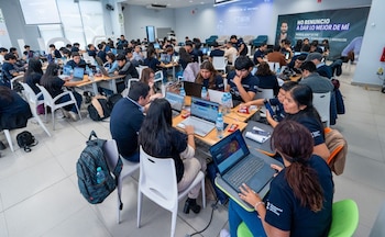 Jóvenes sentados frente a computadoras portátiles en mesas de un salón amplio con pantallas y carteles de fondo
