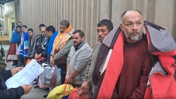 Grupo de hombres en fila, algunos esposados y cubiertos con mantas, mientras una persona sostiene documentos