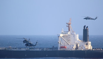 Dos helicópteros militares grises vuelan sobre un buque petrolero oscuro con superestructura blanca en el mar azul, bajo un cielo despejado