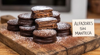 Una pila de alfajores de chocolate con relleno de dulce de leche y azúcar impalpable sobre una tabla de madera, junto a un cartel que dice ALFAJORES SIN MANTECA.
