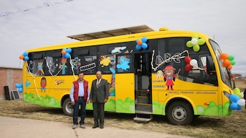 Autobús amarillo con dibujos infantiles, globos y paneles solares. Dos hombres posan delante del vehículo en un terreno abierto con cielo nublado