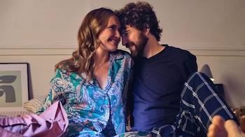 Un hombre con barba y una mujer con pijama estampado sonríen sentados en una cama mientras juntan sus frentes