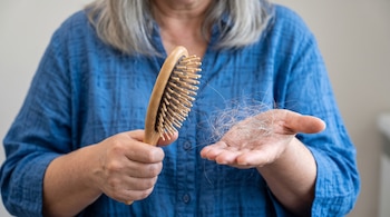 Primer plano de una mujer de mediana edad con cabello canoso y camisa azul, sosteniendo un cepillo de madera con pelo y un mechón de cabello en su palma.
