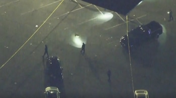 Vista aérea de un estacionamiento oscuro de noche con figuras humanas, vehículos, cinta amarilla de la policía y escombros dispersos tras un incidente