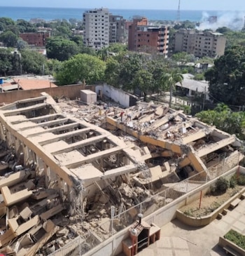Edificio derrumbado con escombros, personas sobre los restos, edificios y árboles al fondo, cielo azul claro, mar a la distancia, humo en el horizonte