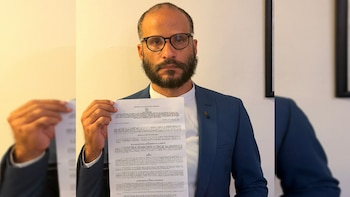 Hombre barbudo con gafas y traje azul sostiene un documento legal blanco frente a un fondo claro. Identificado como Rory Branker