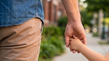 Primer plano de la mano de un adulto sujetando la mano de un niño. El adulto viste camisa de mezclilla y pantalón caqui. Fondo de calle borroso