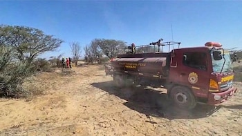 Un camión de bomberos rojo estacionado en un terreno árido, con hombres uniformados al fondo. Hay vegetación seca y un cielo despejado