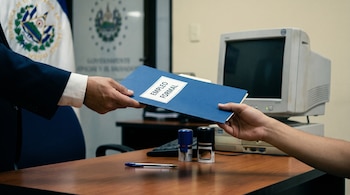 Mano con traje entrega carpeta azul "Empleo Formal" a otra mano. En el escritorio: bolígrafo, sellos y monitor. Fondo desenfocado con escudo de El Salvador.