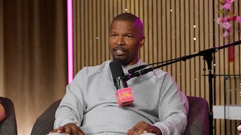Jamie Foxx sentado en un sillón con sudadera gris habla ante un micrófono en un estudio decorado con paneles de madera
