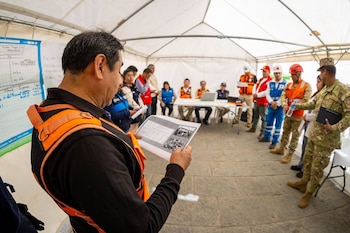Un hombre lee un reporte impreso dentro de una carpa blanca rodeado por socorristas, personal de emergencia y militares