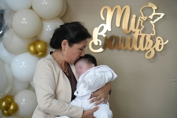 Una mujer besa la frente de un bebé vestido de blanco delante de varios globos y un letrero dorado con la frase Mi Bautizo